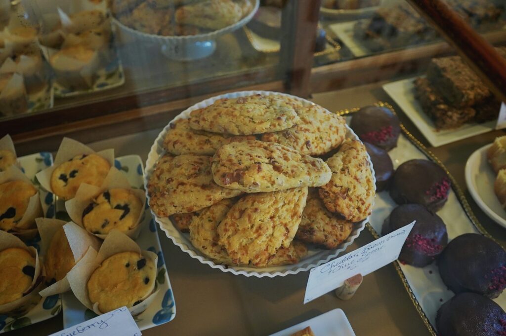 Bakery display case with a plate of cheddar scones, blueberry muffins and chocolate-dipped cookies