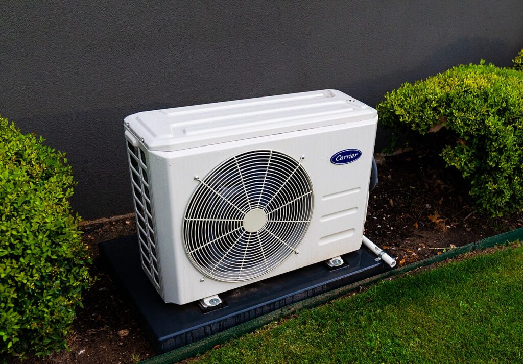 Outdoor heat pump unit on a pad beside a house, with shrubs on both sides