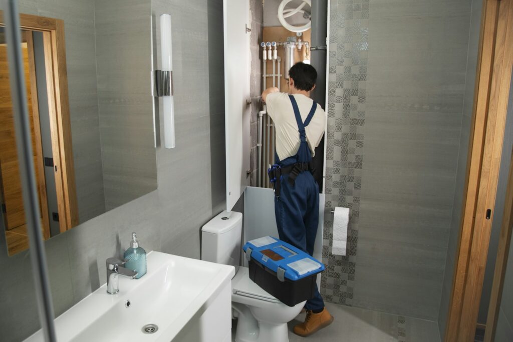 Plumber in overalls working on pipes behind an open access panel in a bathroom, toolbox on the toilet